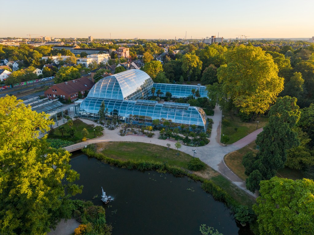 Luftaufnahme der Flora in Köln mit Glashaus, Weiher und umgebender Parkanlage.