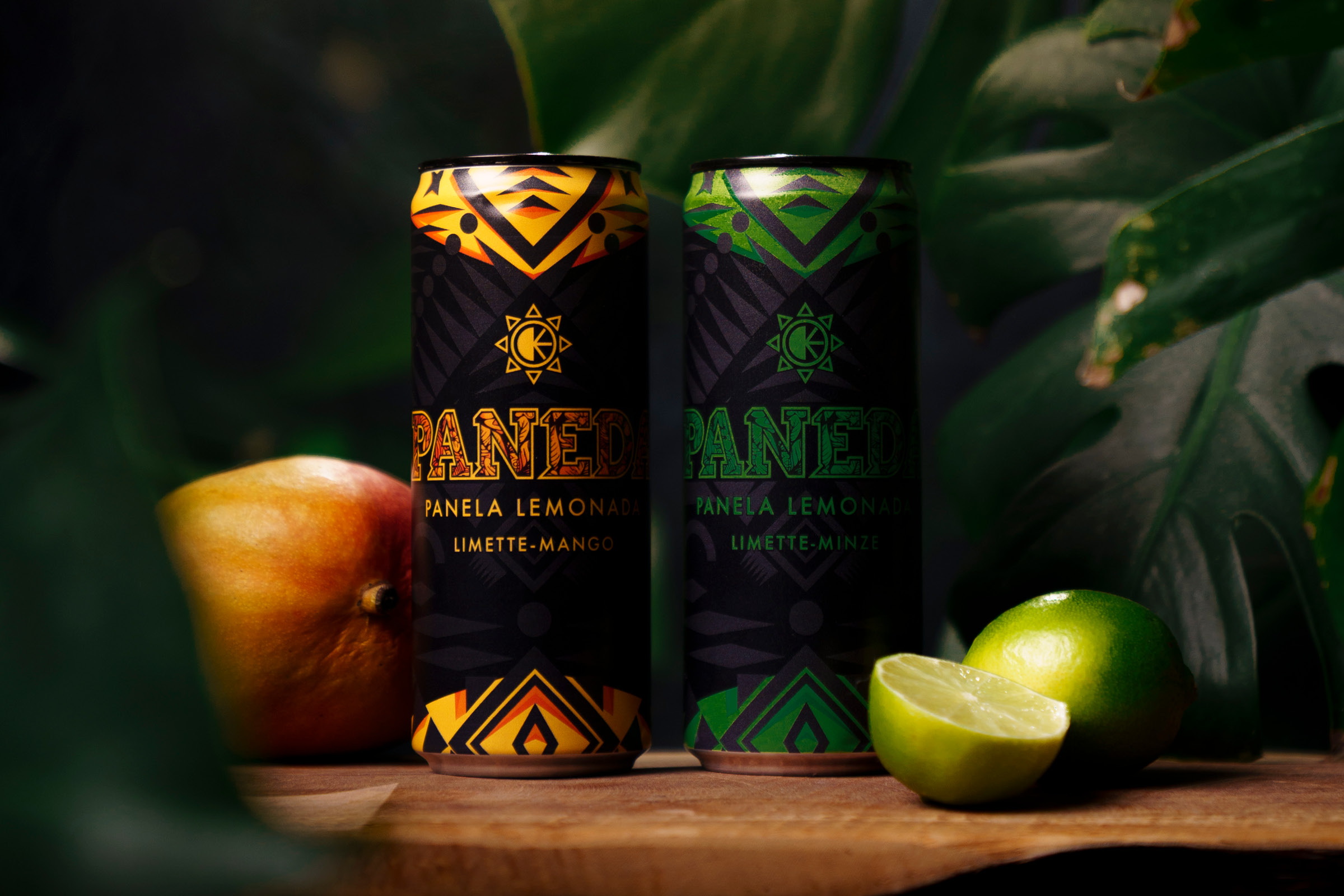 Zwei Dosen Paneda Panela Lemonada in Limette-Mango und Limette-Minze auf Holz, daneben Mango und aufgeschnittene Limette vor dunklem Blattwerk.