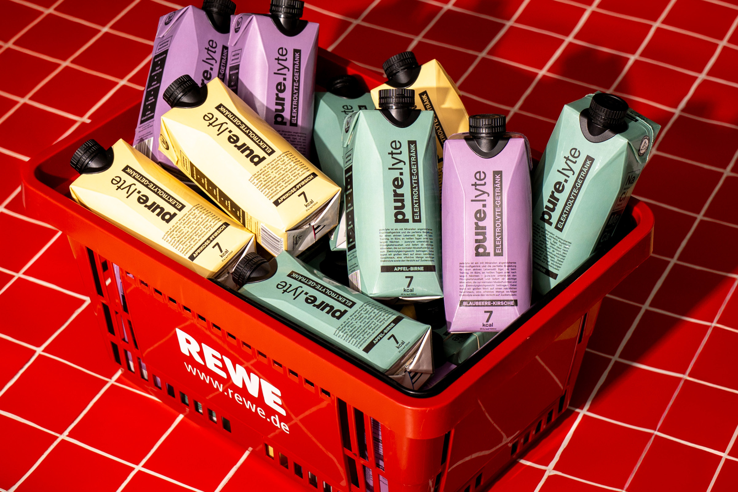 Elektrolyt-Getränke von pure.lyte in mehreren Sorten in einem roten REWE-Einkaufskorb auf rotem Fliesenboden.